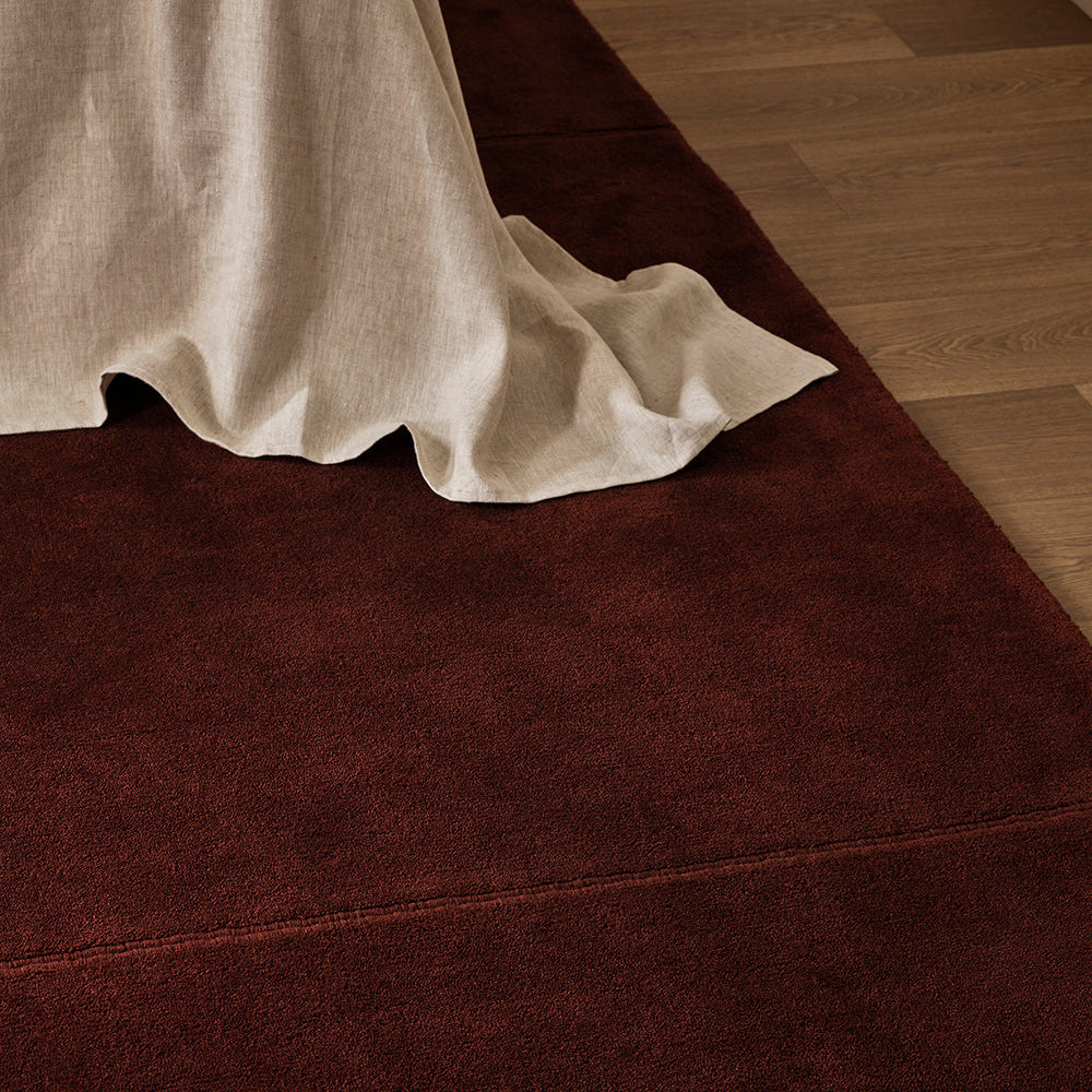 A dark reddish-brown area rug partially shown under a draped off-white linen throw on a light wood floor.