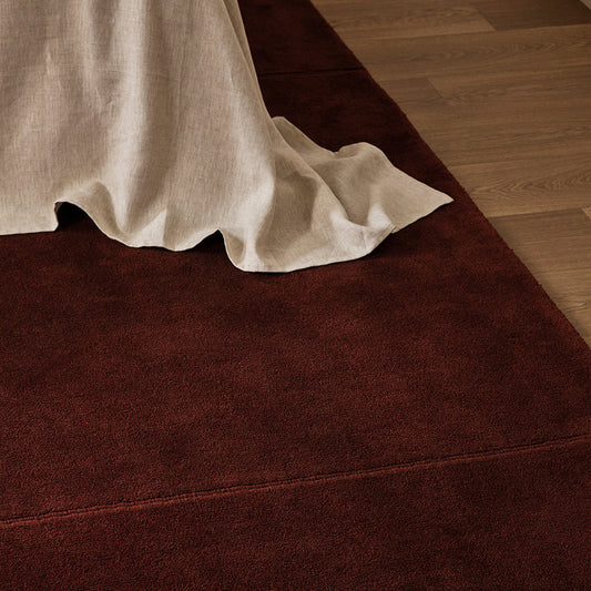 A dark reddish-brown area rug partially shown under a draped off-white linen throw on a light wood floor.