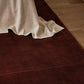 A dark reddish-brown area rug partially shown under a draped off-white linen throw on a light wood floor.