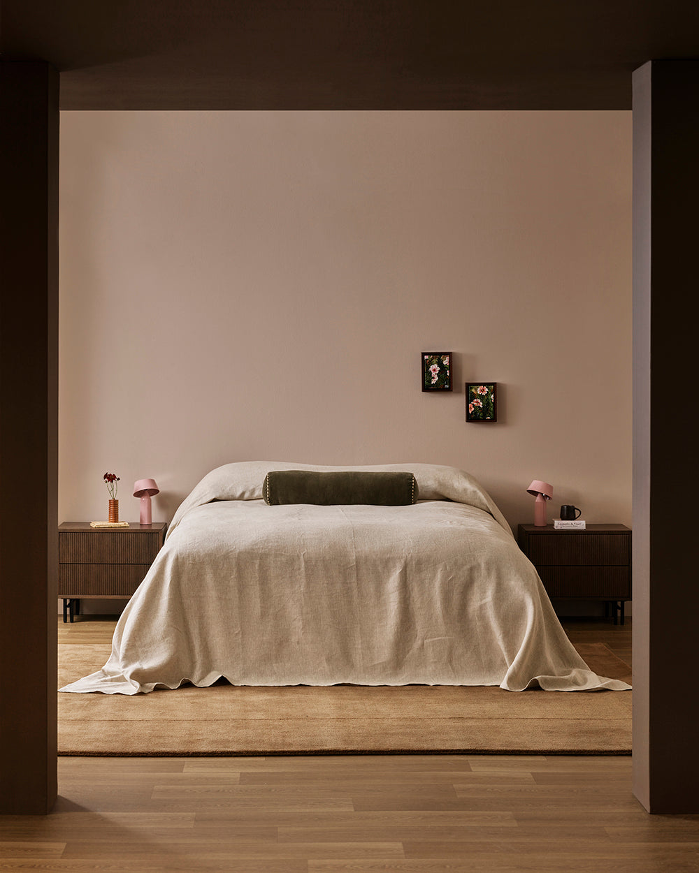A minimalist bedroom scene viewed through a dark doorway, featuring a neatly made bed with cream bedding and a dark olive bolster pillow against a warm beige wall, with matching bedside tables and pink lamps on both sides.