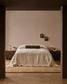 A minimalist bedroom scene viewed through a dark doorway, featuring a neatly made bed with cream bedding and a dark olive bolster pillow against a warm beige wall, with matching bedside tables and pink lamps on both sides.