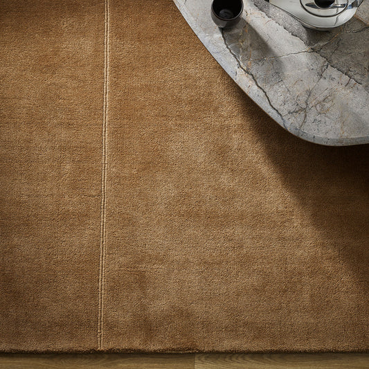A close-up of a warm brown textured rug with a single vertical seam, shown in a styled interior setting with a marble coffee table and a teapot.
