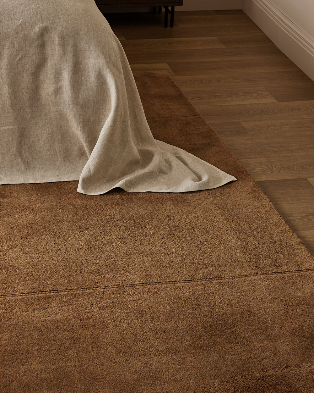 A brown textured area rug styled in a bedroom setting, partially covered by a light beige linen throw blanket on a wooden floor.