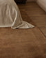 A brown textured area rug styled in a bedroom setting, partially covered by a light beige linen throw blanket on a wooden floor.