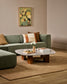 A styled living room scene featuring a sage green sectional sofa with neutral and earthy throw pillows, a marble-top coffee table with wooden legs, a large beige rug, and a framed abstract floral painting on the wall.