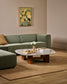A styled living room scene featuring a sage green sectional sofa, a matching ottoman, a marble-top coffee table with wooden legs, a beige rug, and a framed abstract floral painting on the wall.