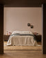 A minimalist bedroom scene viewed through an open doorway, featuring a neatly made bed with cream bedding and a gray throw, flanked by dark wood nightstands with pink lamps against a warm beige wall.