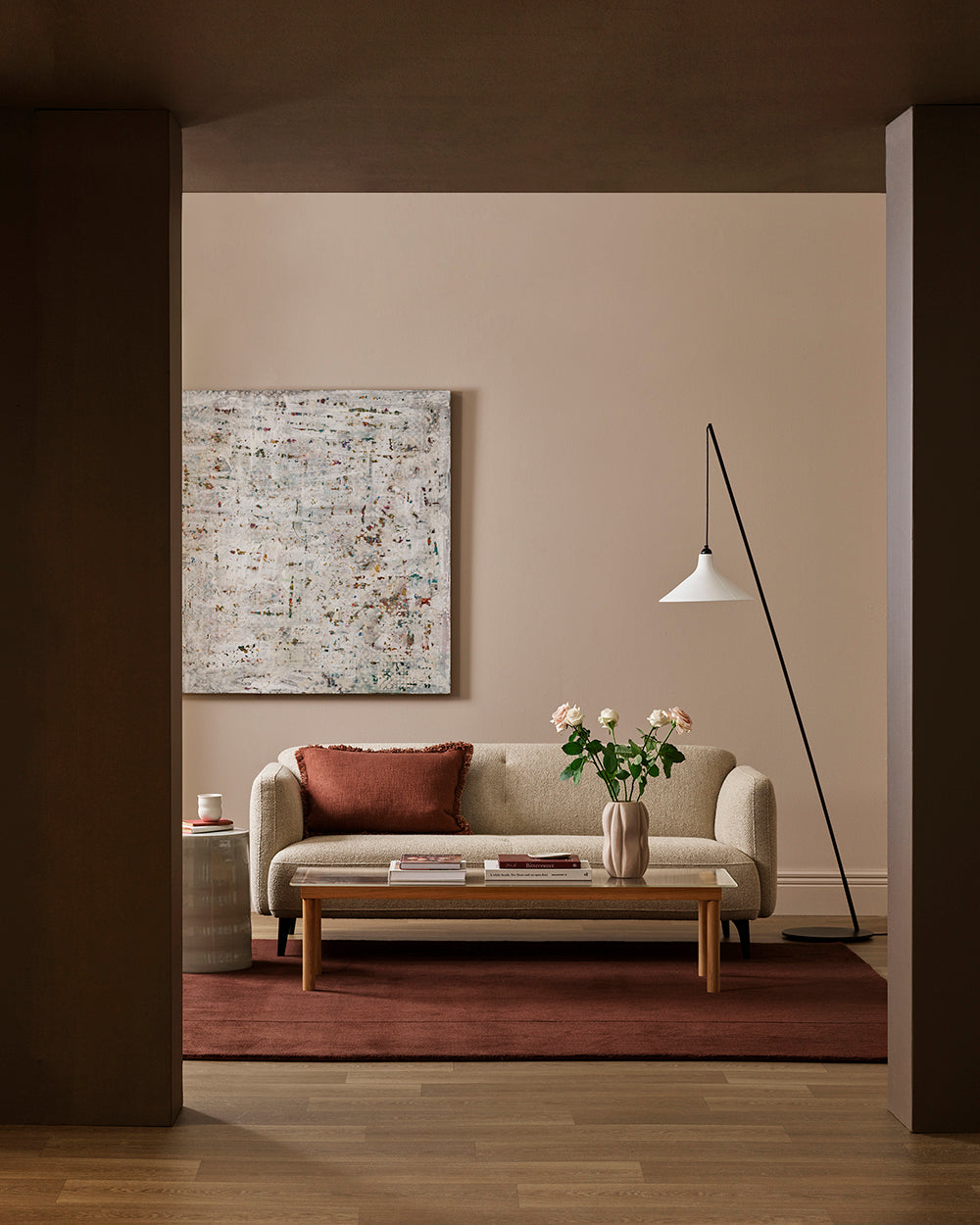 A styled living room interior featuring a beige sofa with a rust red pillow, a wooden coffee table, a red rug, a black floor lamp, and an abstract canvas painting on a warm beige wall.