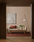 A styled living room interior featuring a beige sofa with a rust red pillow, a wooden coffee table, a red rug, a black floor lamp, and an abstract canvas painting on a warm beige wall.