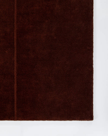 A close-up of a dark red textured wool rug, showing its soft, nubby surface and a stitched edge, set against a plain white background.