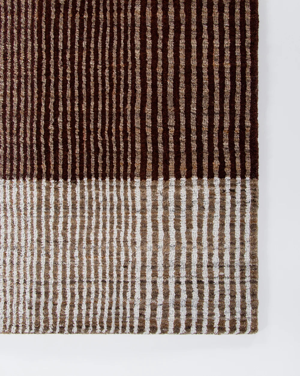 A close-up of a woven textile or rug corner showing a striped pattern with a dark brown upper section and a lighter beige-and-white lower section, photographed against a plain light background.