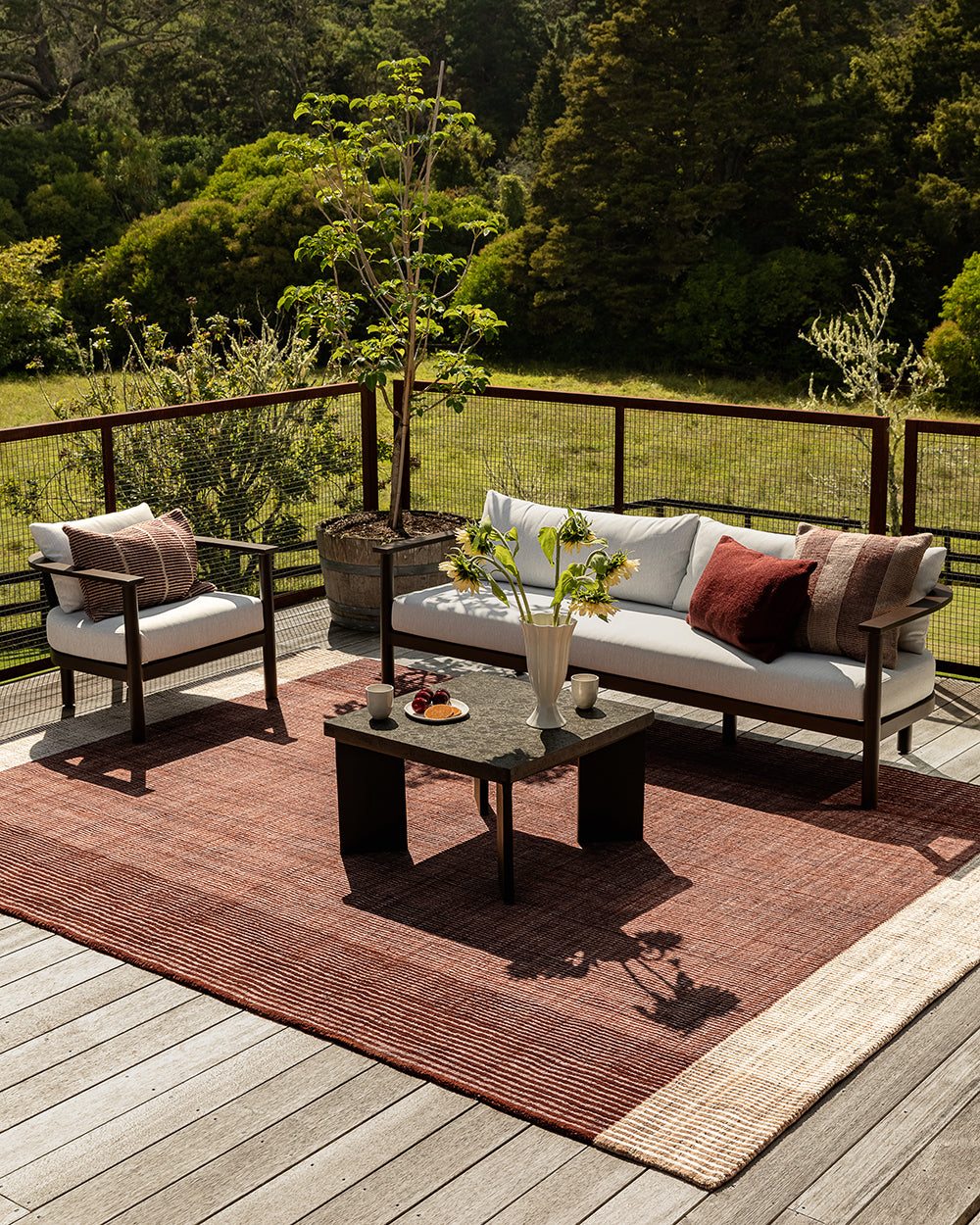 Outdoor patio furniture set on a wooden deck with a rust-red woven area rug, a white sofa with white and red cushions, a matching armchair, and a dark coffee table, with a lush green forest background.