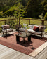 Outdoor patio furniture set on a wooden deck with a rust-red woven area rug, a white sofa with white and red cushions, a matching armchair, and a dark coffee table, with a lush green forest background.