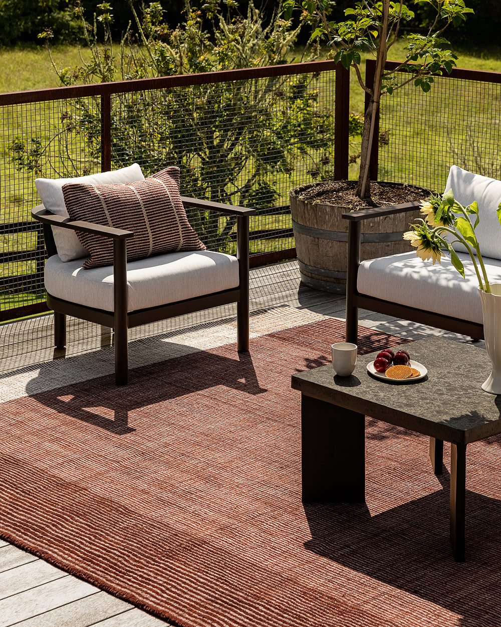 A styled outdoor patio scene featuring a rust-brown textured area rug, a dark wood armchair with white cushions, a matching side chair, and a low stone coffee table on a deck with a railing and greenery in the background.