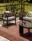 A styled outdoor patio scene featuring a rust-brown textured area rug, a dark wood armchair with white cushions, a matching side chair, and a low stone coffee table on a deck with a railing and greenery in the background.