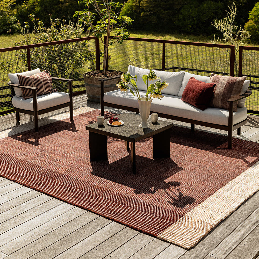 Outdoor patio furniture set on a wooden deck with a rust-red woven area rug, a white sofa with white and red cushions, a matching armchair, and a dark coffee table, with a lush green forest background.