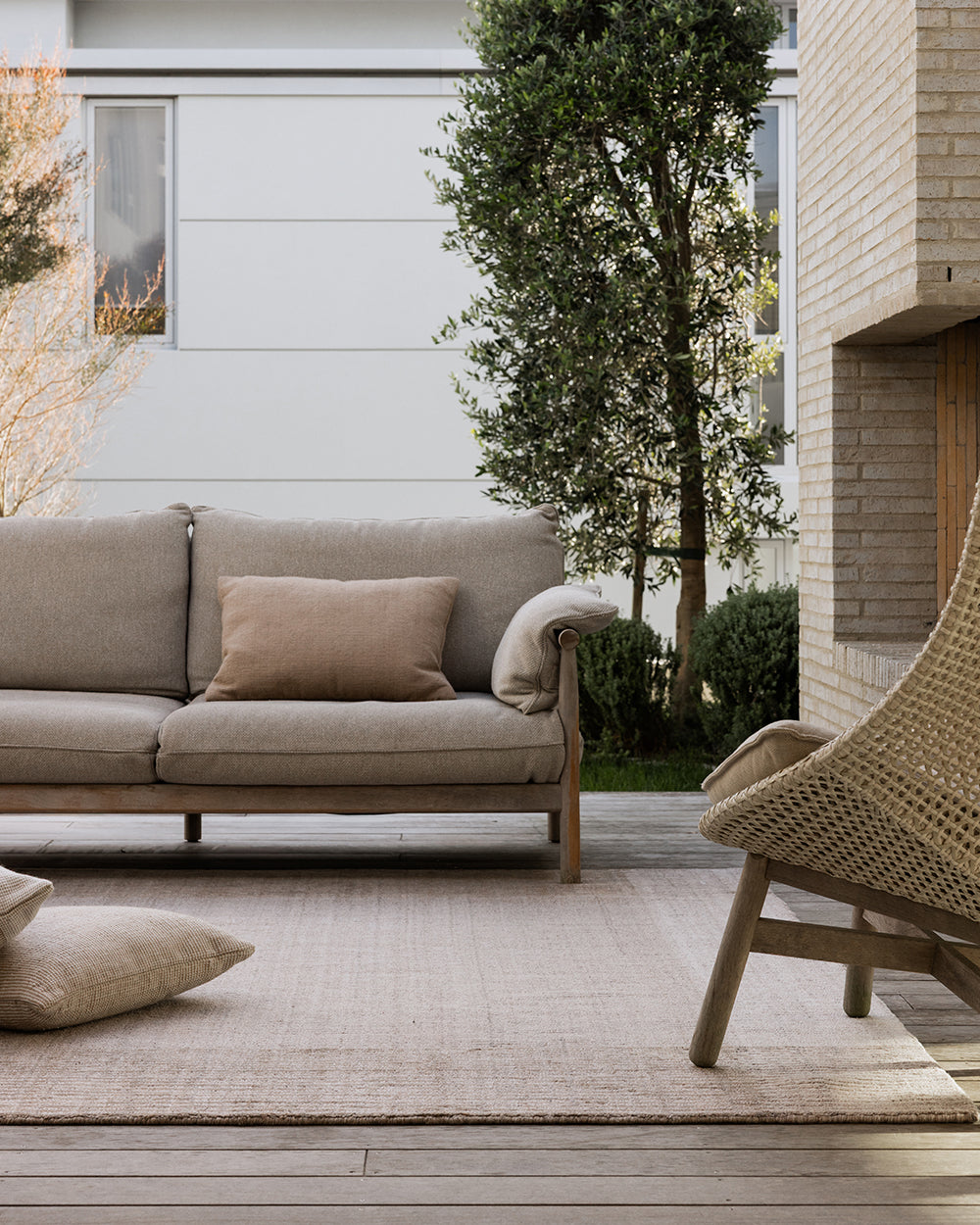 A modern outdoor living room scene featuring a light beige fabric sofa with a matching throw pillow, a woven rattan armchair, a neutral rug, and a brick fireplace in the background.