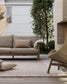 A modern outdoor living room scene featuring a light beige fabric sofa with a matching throw pillow, a woven rattan armchair, a neutral rug, and a brick fireplace in the background.