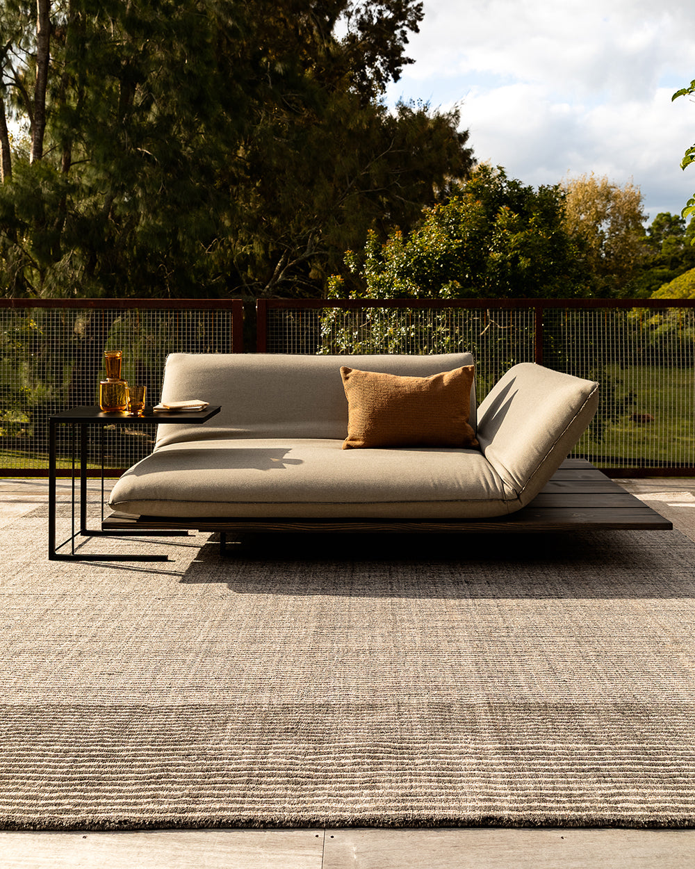 A modern outdoor daybed with a light beige cushion and a brown accent pillow, set on a textured gray rug with a black metal side table. The background features a wire mesh fence and lush green trees under a cloudy sky.