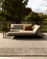 A modern outdoor daybed with a light beige cushion and a brown accent pillow, set on a textured gray rug with a black metal side table. The background features a wire mesh fence and lush green trees under a cloudy sky.