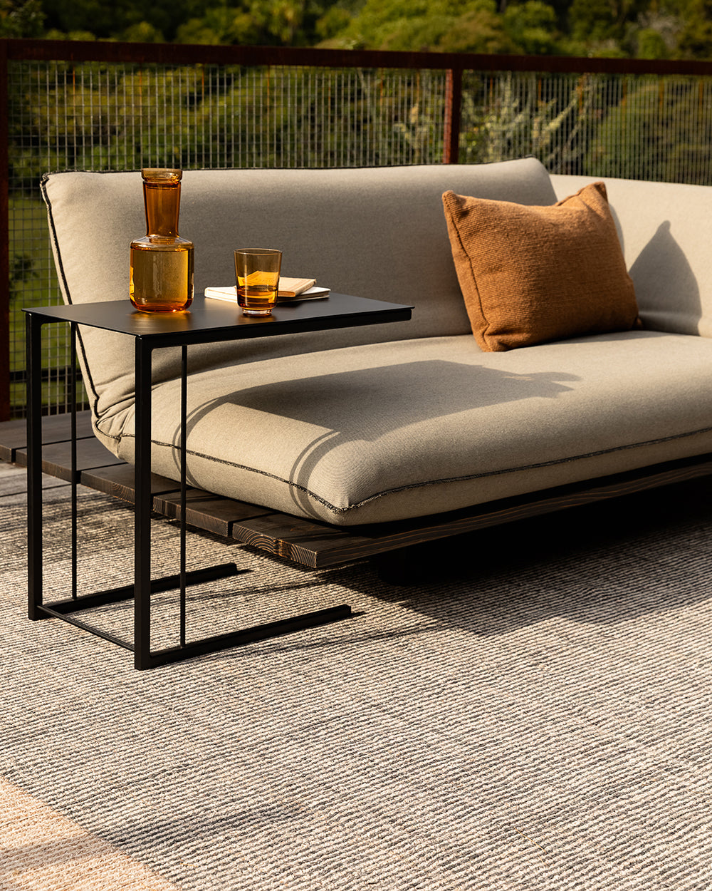 Outdoor patio furniture set featuring a beige cushioned daybed with a black metal side table, styled with amber glass tumblers and a brown throw pillow on a textured rug.