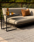 Outdoor patio furniture set featuring a beige cushioned daybed with a black metal side table, styled with amber glass tumblers and a brown throw pillow on a textured rug.