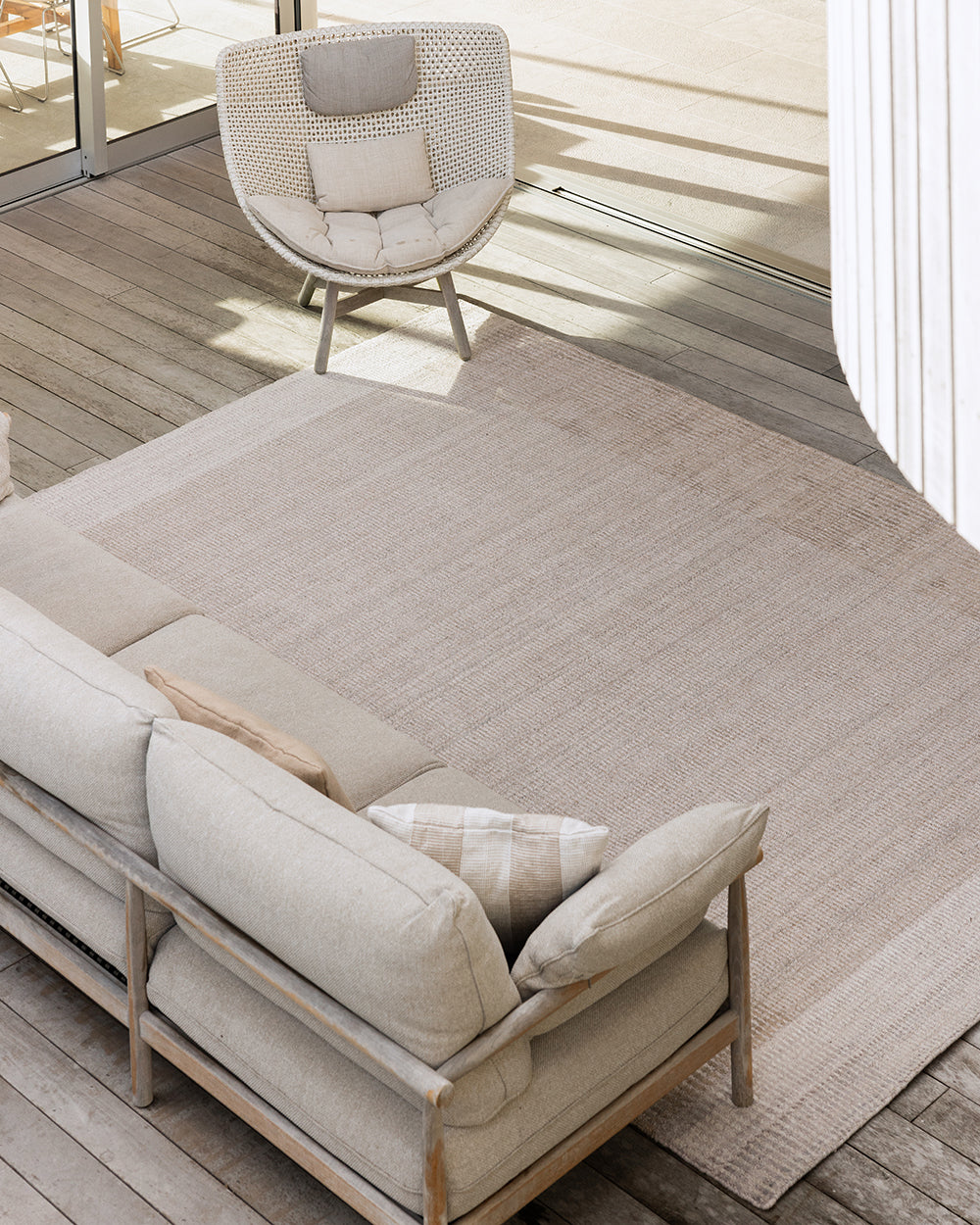 A large rectangular outdoor area rug in a light beige tone, shown on a wooden deck beside a beige sofa and a woven accent chair.