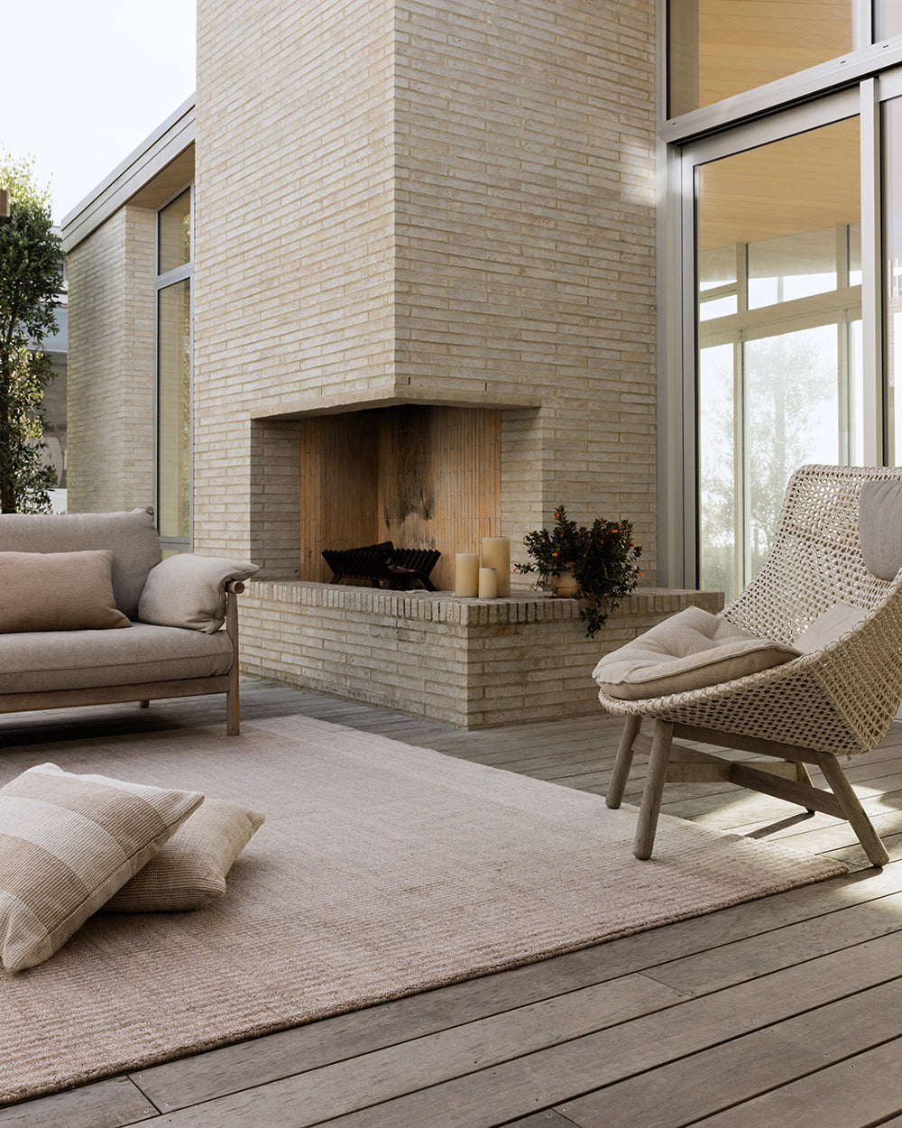 A modern outdoor patio scene featuring a light beige woven armchair with a cushion, a beige sofa, a large textured area rug, and a built-in brick fireplace in a contemporary house.