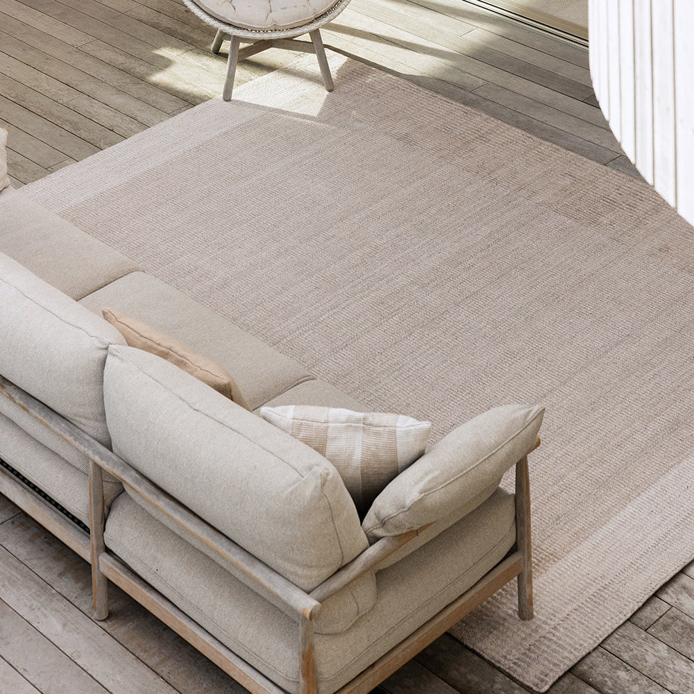 A large rectangular outdoor area rug in a light beige tone, shown on a wooden deck beside a beige sofa and a woven accent chair.
