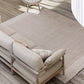 A large rectangular outdoor area rug in a light beige tone, shown on a wooden deck beside a beige sofa and a woven accent chair.