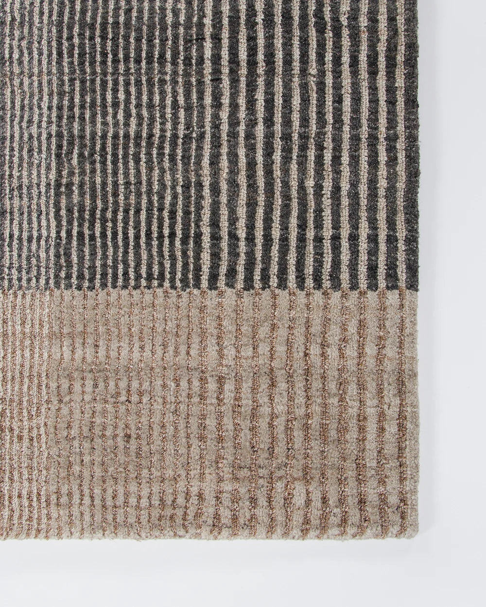Close-up of a textured woven rug corner showing a striped pattern in dark charcoal gray and warm beige on a white background.