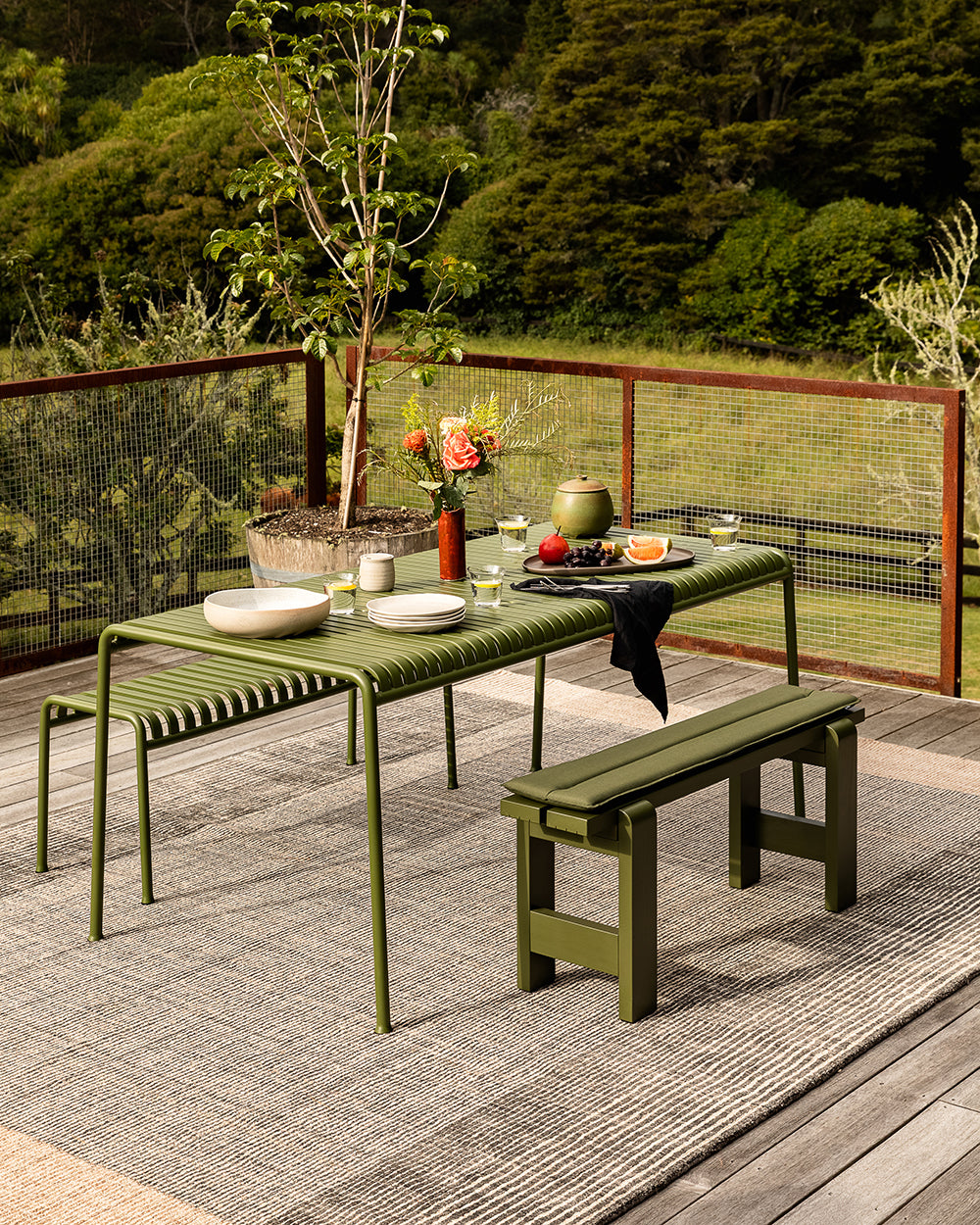 An outdoor dining set consisting of a rectangular table and two matching benches in olive green, styled on a gray textured rug with a wooden deck and lush green garden in the background.