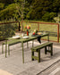 An outdoor dining set consisting of a rectangular table and two matching benches in olive green, styled on a gray textured rug with a wooden deck and lush green garden in the background.