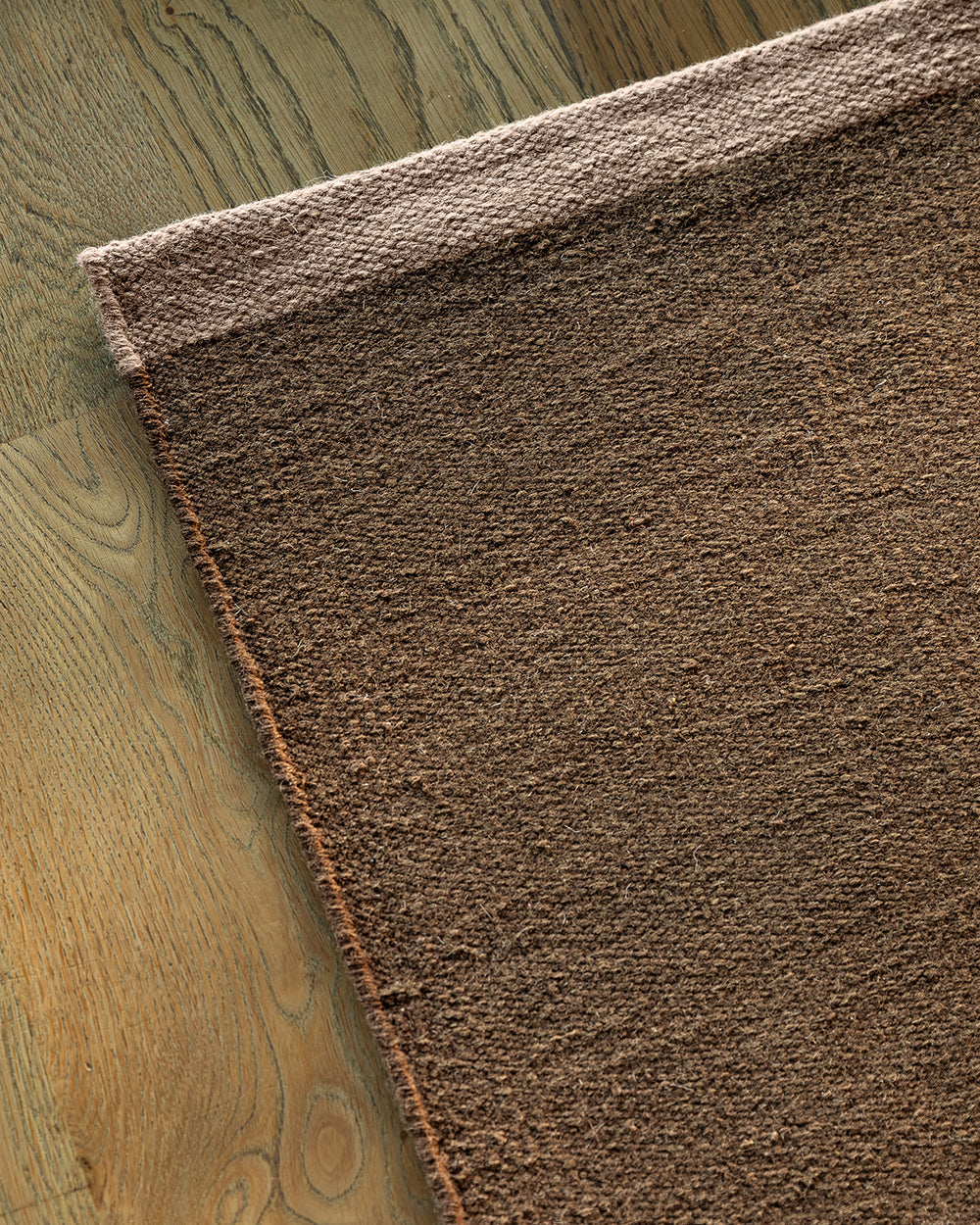 A close-up of a brown textured area rug with a fringed edge, laid out on a light brown wooden floor.