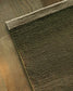 A close-up of a textured area rug corner with a thick beige braided border, laid on a medium brown wood floor.