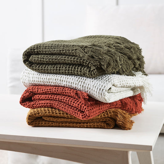 Stack of cotton waffle throws folded. Olvie, Ecru, Brick and Toffee colours sitting on a white table