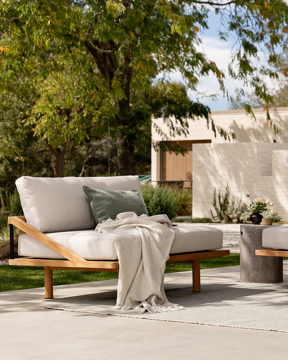 Outdoor patio setting with a wooden sofa, cushions, and a blanket in a garden.