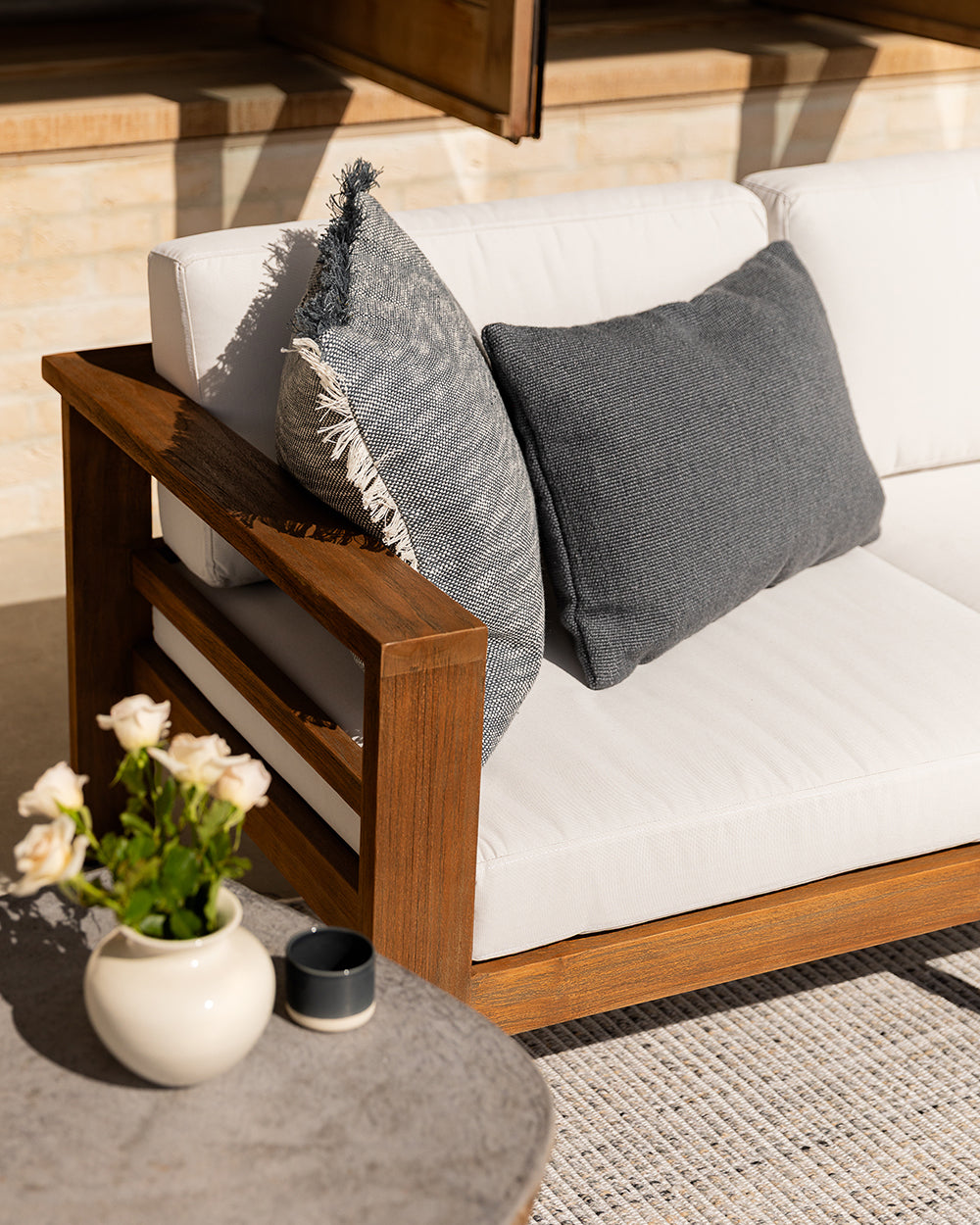 White outdoor sofa with wooden armrest and gray cushions, accompanied by a small table with flowers and a candle.