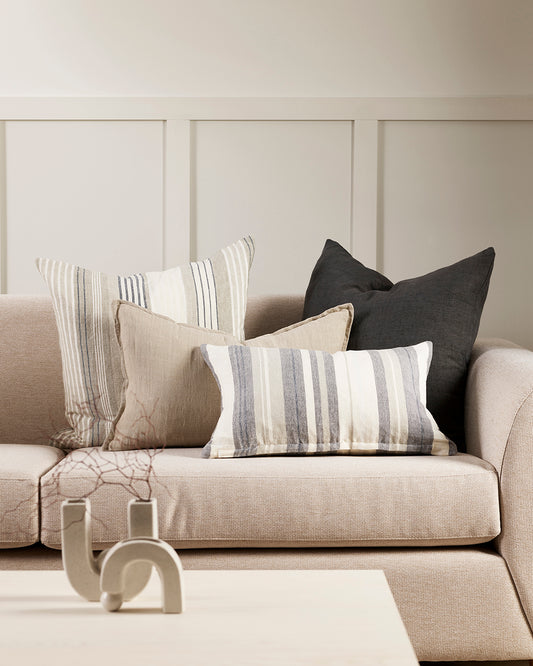 Lifestyle shot of a group of cushions in greys, charcoals and creams, some plain, some striped on a cream modern sofa. Behind the sofa is a grey wainscotting wall and a white coffee table with a white curved candlestick style vase and dried botanics in