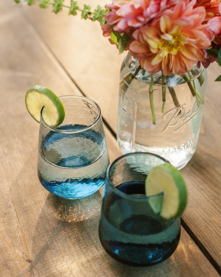 Two glasses with blue drinks and lime slices on a wooden table, with a Mason jar of flowers in the background.