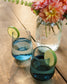 Two glasses with blue drinks and lime slices on a wooden table, with a Mason jar of flowers in the background.