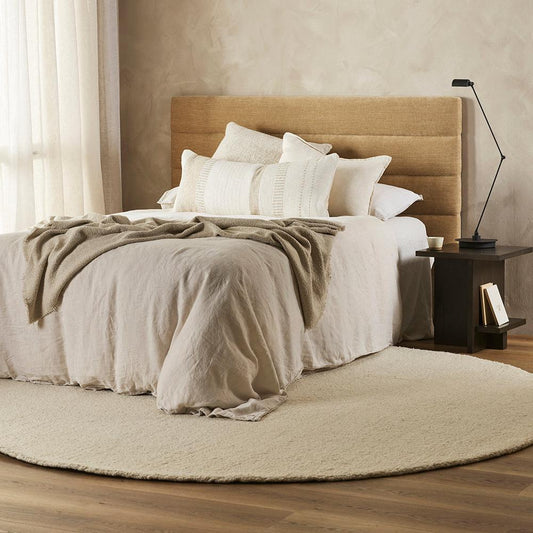 white boucle wool round rug with a beige padded  headboard on a bed covered with white and grey linen. Wooden floors with linen curtains with light shading through
