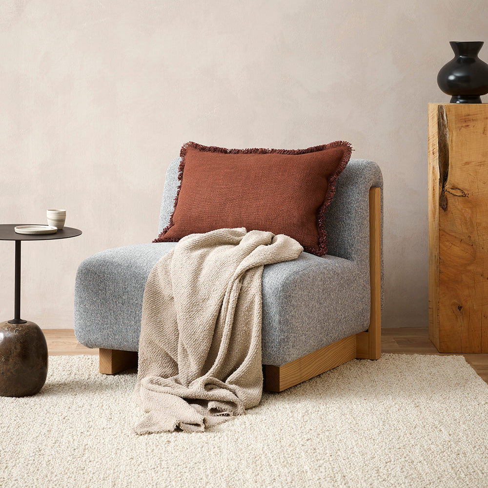 A gray cushion with a chunky textural weave and soft fringing, placed on a blue armchair in a room with a wooden side table and a round mirror.