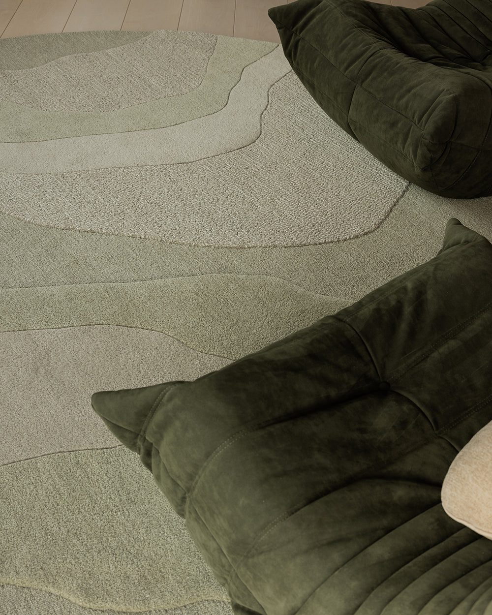 A close-up of a textured area rug with organic wavy lines in light sage green and cream, styled with dark olive green floor cushions.