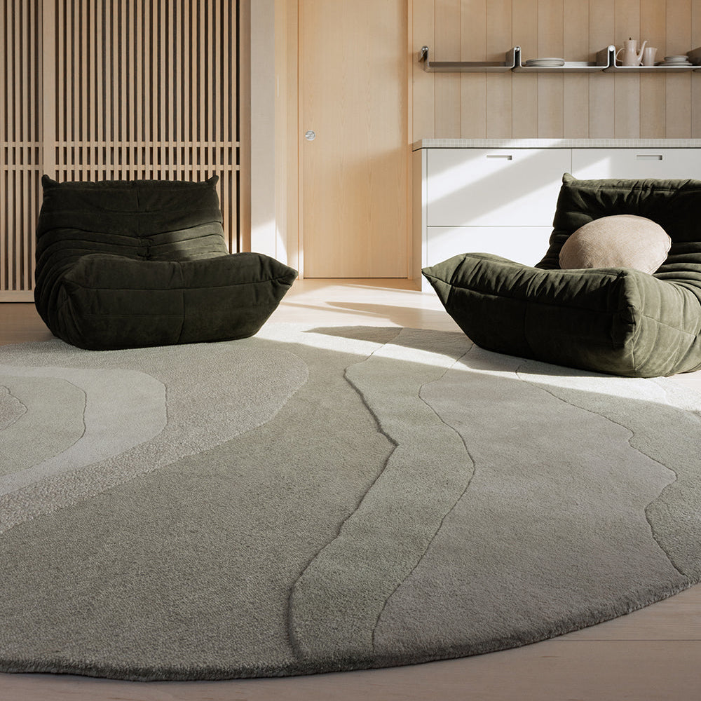 A modern living room scene featuring a large, abstract-patterned gray area rug on the floor, with two dark olive green velvet lounge chairs placed on either side of the room. The background consists of light wood-paneled walls and ceiling, a wooden slatted screen, and a white cabinet.