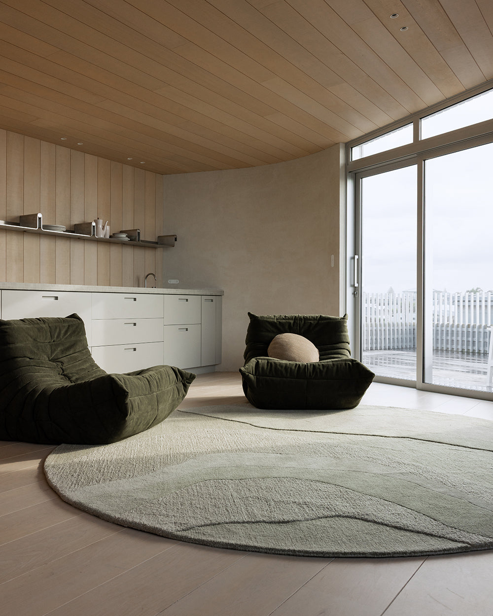 A modern living room interior with two dark olive green lounge chairs placed on a large round light gray rug. The room has light wood flooring and ceiling, a white cabinet along the left wall, and large floor-to-ceiling windows on the right.