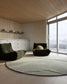 A modern living room interior with two dark olive green lounge chairs placed on a large round light gray rug. The room has light wood flooring and ceiling, a white cabinet along the left wall, and large floor-to-ceiling windows on the right.