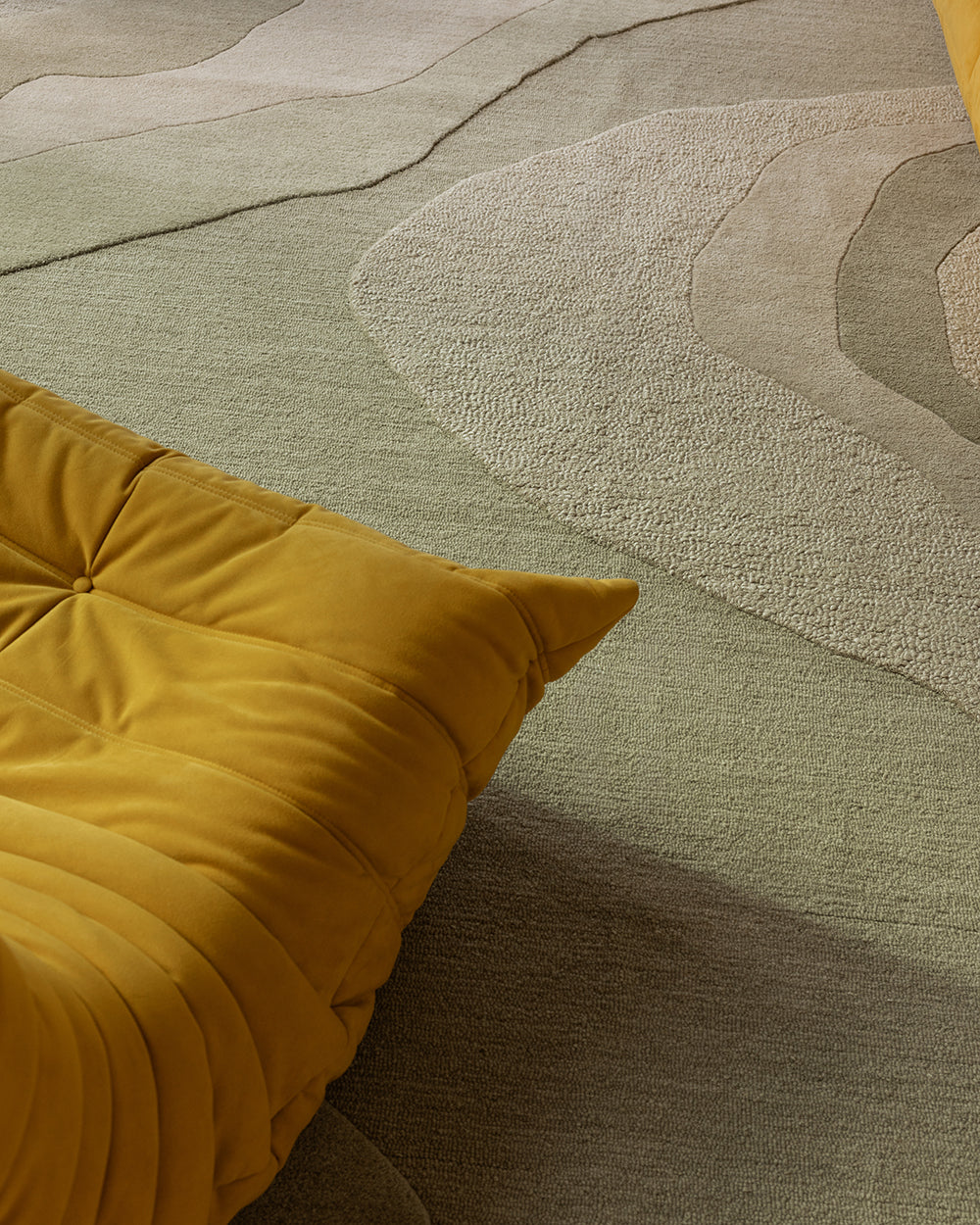 A close-up of a modern area rug with layered, organic wavy patterns in muted sage green, taupe, and gray, styled with a mustard yellow tufted cushion in the foreground.