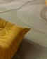 A close-up of a modern area rug with layered, organic wavy patterns in muted sage green, taupe, and gray, styled with a mustard yellow tufted cushion in the foreground.