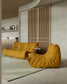 A modern living room scene featuring a mustard yellow tufted lounge chair in the foreground and a matching low sofa in the background, set on a light rug in front of a slatted wooden cabinet.